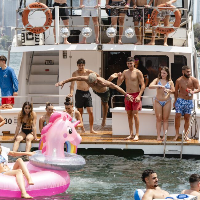 A group of people enjoying a sunny day on a boat. Some are swimming, and others are relaxing on inflatable floats, including a pink unicorn. The city skyline and trees are visible in the background.