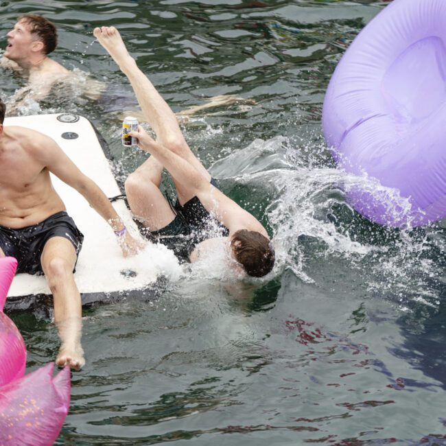 Three men are playing in the water. Two are on an inflatable platform, with one laughing and holding a can. The other is falling backward into the water, causing a splash. There are colorful inflatable floats nearby.