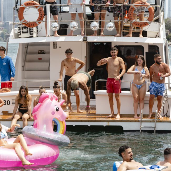 A group of young adults in swimsuits enjoy a sunny day on a boat. Some are on the deck, while others are in the water with inflatable floats, including a pink unicorn. The city skyline and greenery are visible in the background.