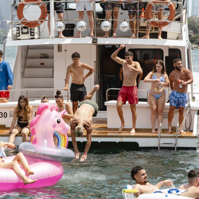 A group of people enjoying a sunny day on a boat. Some are lounging on inflatable floats, others swim nearby. The boat is docked with more people on board. The scene is lively and cheerful, set against a backdrop of an urban skyline and water.