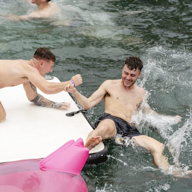 Two men are having fun in a body of water. One man, sitting on a floating platform, reaches to help another man in the water. A bright pink inflatable is in the foreground, and others are swimming nearby.