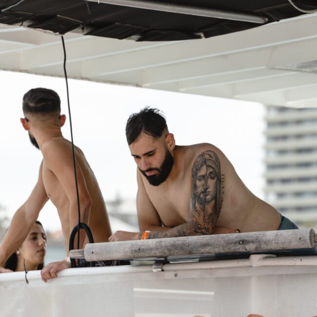 A man with tattoos leans on a boat railing, looking down. Another man stands nearby, facing away. A woman is partially visible in the background. The boat banner reads "BAREFOOT" with the ocean and a building in the background.