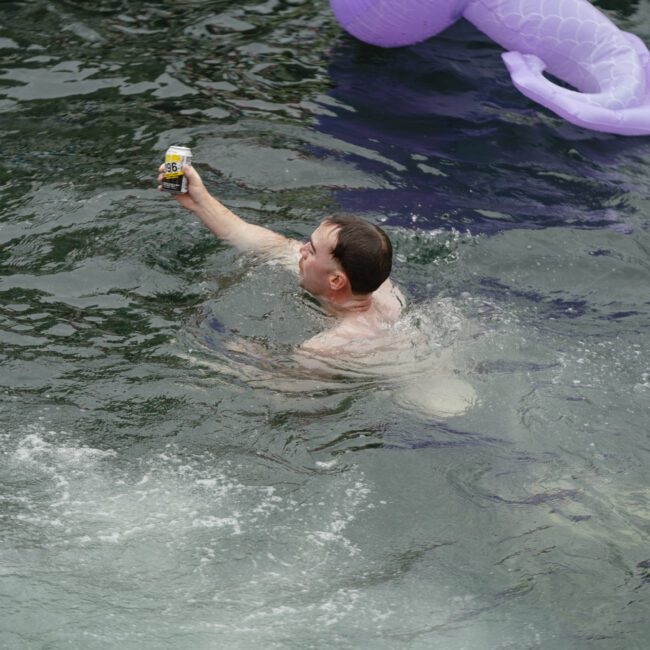 A person is swimming in a body of water, holding a can of drink above the surface. There are inflatable toys floating nearby, including a purple one shaped like a creature's tail. The water shows some ripples around the swimmer.