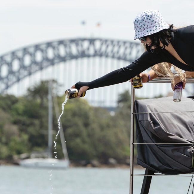 A person wearing a bucket hat and long sleeves is on a boat, pouring a drink overboard. In the background, a large bridge spans across the water, and trees line the shore. The scene suggests a casual outdoor setting.