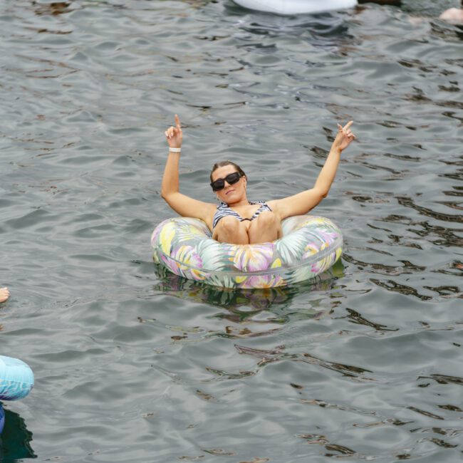 A person wearing sunglasses relaxes on an inflatable tube in the water, smiling and making peace signs with both hands. Other people on inflatables are nearby, enjoying the sunny day.