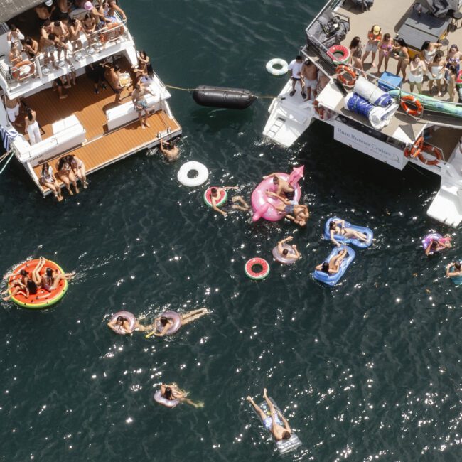 Aerial view of people enjoying a sunny day on a lake with two boats docked close together. People are swimming, lounging on inflatable floats, and socializing on the boats and in the water.