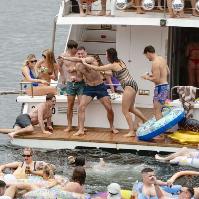 A group of people on a boat enjoying a party. Some are dancing and playing on the deck, while others relax on colorful inflatable tubes in the water. There are drinks and laughter, creating a lively, fun atmosphere.