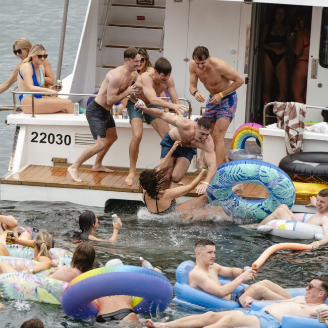 A lively scene on a boat with people having fun; some are helping a woman who is falling into the water surrounded by colorful inflatable rings. Several others are already in the water, enjoying the sunny day.