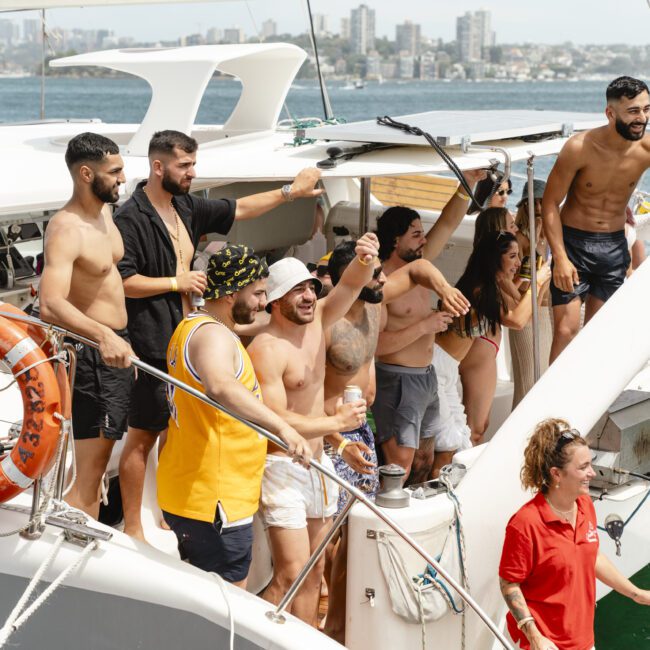 A group of people on a boat, some in swimwear, others carrying drinks, are smiling and enjoying a sunny day. A cityscape is visible in the background. A crew member in a red shirt stands nearby.