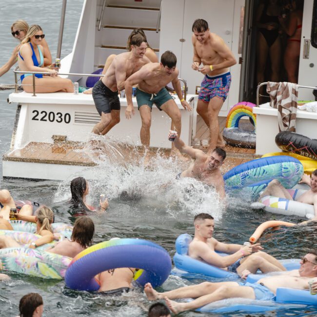 People enjoy a lively boat party, with some swimming and others lounging on inflatable floats in the water. Several individuals are standing and mingling on the boat, with a festive atmosphere.