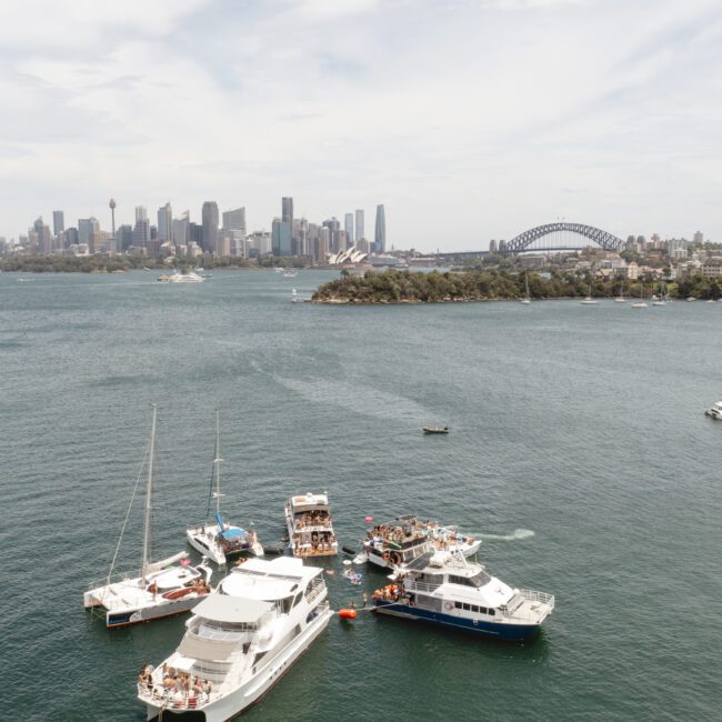 Aerial view of several boats gathered on a body of water with a city skyline and iconic bridge in the background under a partly cloudy sky.