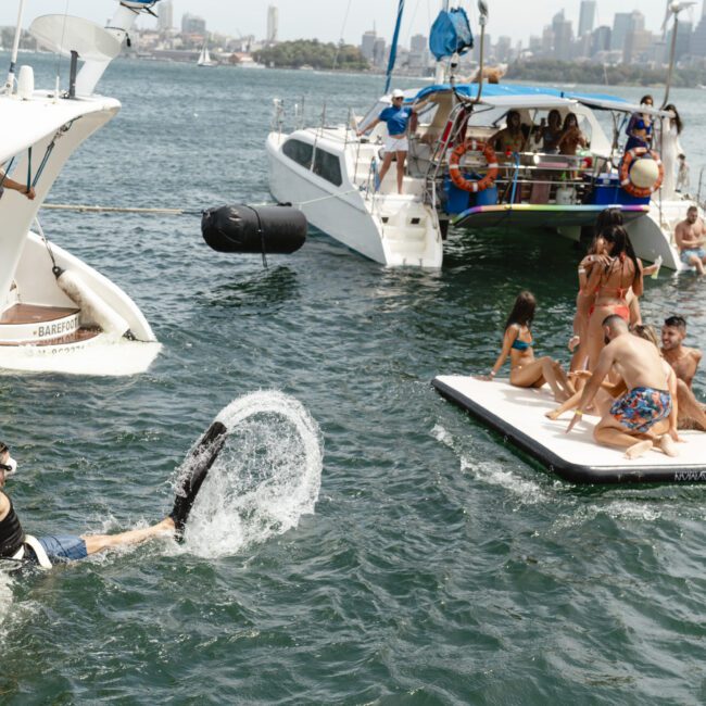 People enjoy a sunny day on the water near several boats. Some are on an inflatable platform, others swim nearby. One person sits at the edge of a boat, while someone dives in, creating a splash. The city skyline is visible in the background.