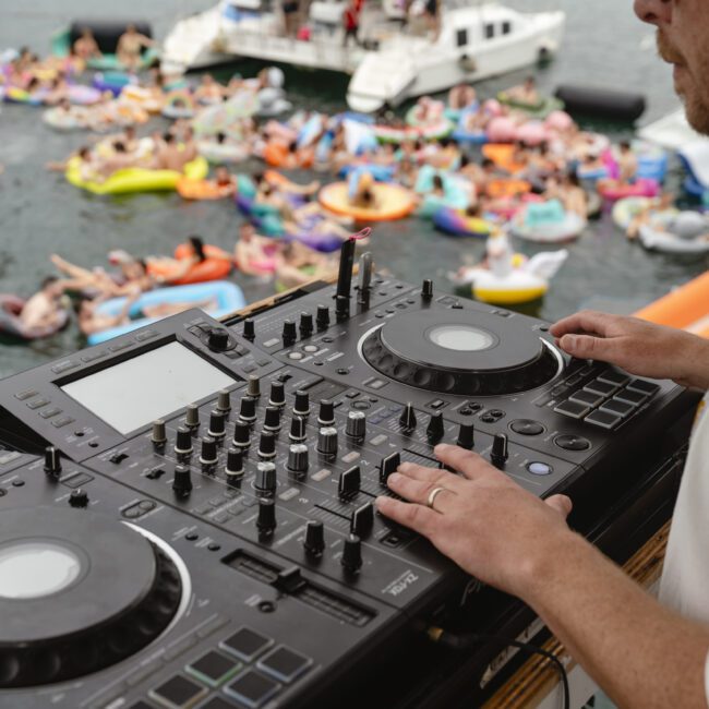 A DJ is playing music on a boat with a mixer and turntables. In the background, people are floating in colorful inflatable tubes on the water, enjoying a party atmosphere.