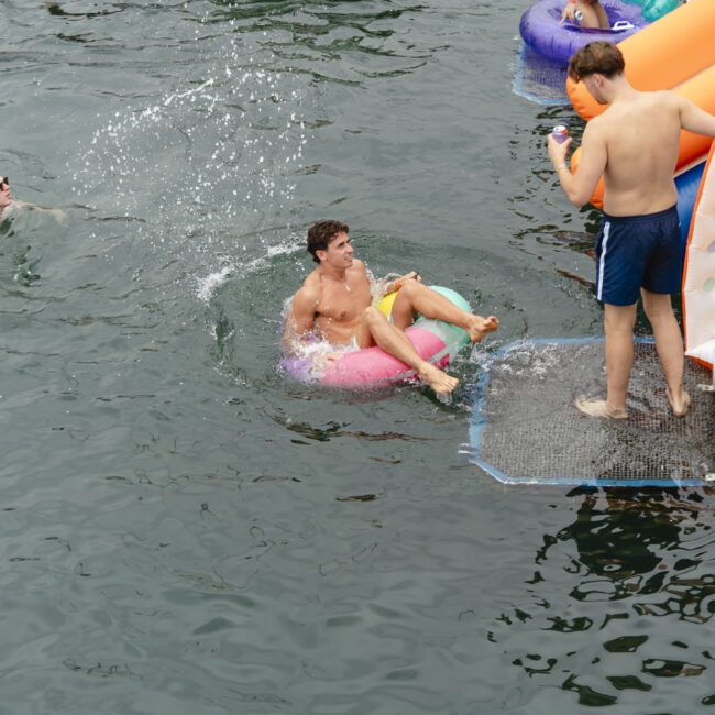 Three people enjoy swimming in a lake. One person floats on a pink inflatable ring, another stands on a platform holding an inflatable mattress, and a third person is swimming nearby, causing splashes in the water.