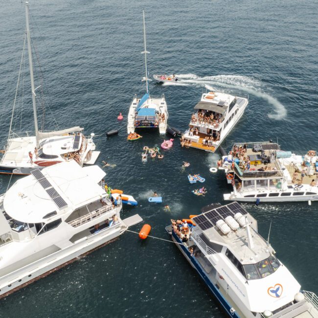 Several boats anchored close together in the ocean, with people swimming and floating on inflatables between them. The scene is lively, with a variety of colorful floats and people enjoying the water.