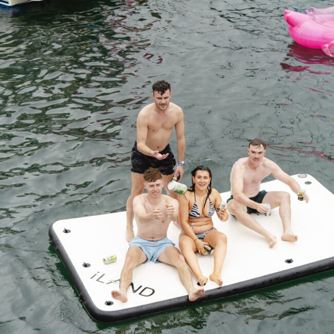 Four people sit on a floating platform in a body of water. They are wearing swimwear, holding drinks, and smiling at the camera. A pink inflatable float is visible nearby.