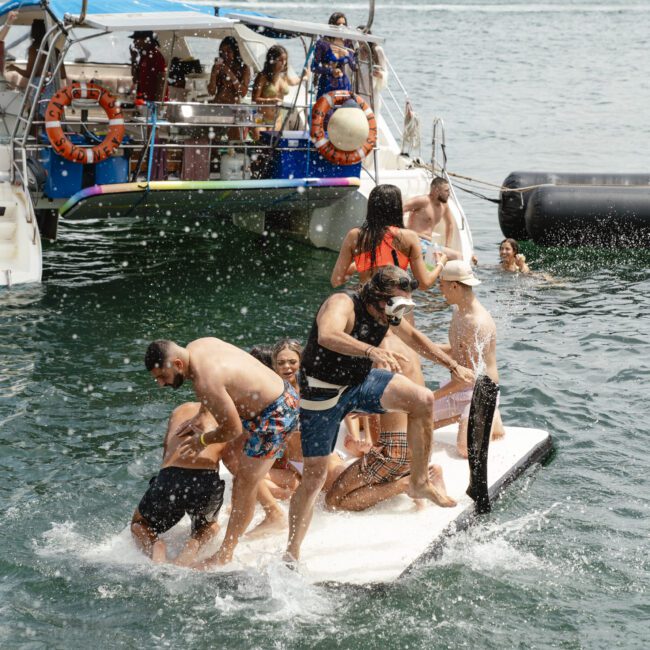 A group of people enjoys splashing and playing on a floating mat beside a boat in a harbor. The city skyline is visible in the background. The atmosphere is lively and fun.