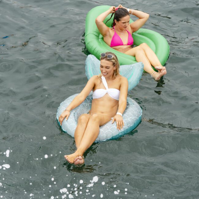 Two women relaxing on inflatable floats in a body of water. One wears a white bikini on a blue float, while the other is in a pink swimsuit on a green float. Splashes of water are in the foreground.