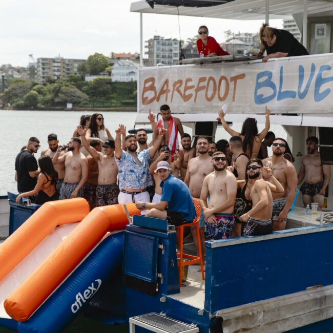 A group of people enjoying a boat party. Some are standing on the deck, while others are in swimwear near orange slides. A banner reads "Barefoot Blue." Buildings and trees are visible in the background.