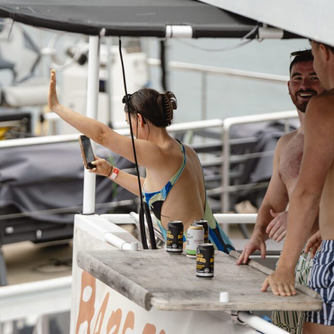 Three people on a boat deck; a woman takes a selfie while two men chat. One man is shirtless, wearing striped shorts. There are drinks on a wooden table. Another boat with floats and water visible in the background.