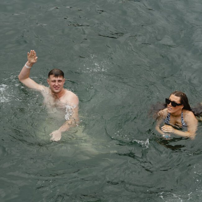 A man and woman are swimming in open water. The man is waving, and the woman is wearing sunglasses. They appear relaxed and enjoying their time in the water.