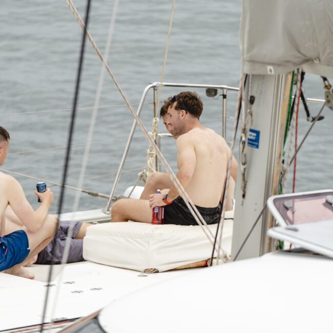 Two men sitting shirtless on a sailboat deck, one holding a drink can. They are facing each other and appear relaxed, with the sea visible in the background. Towels and other boating equipment are around them.