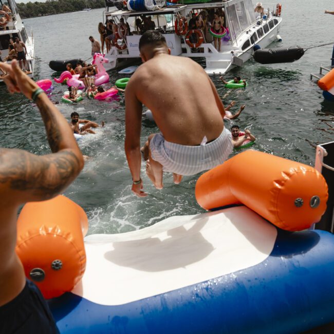 A group of people enjoy a lively boat party on a lake. One person is jumping off an inflatable, while others relax on flotation devices. Boats are anchored nearby, and the atmosphere is festive and fun.