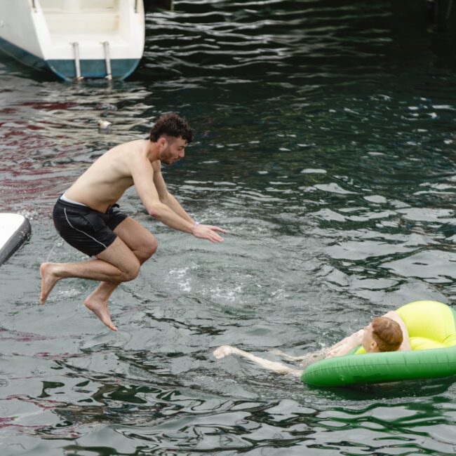 A man in black swim trunks jumps off a platform into the water where another person floats on a green inflatable ring. Boats are visible in the background.