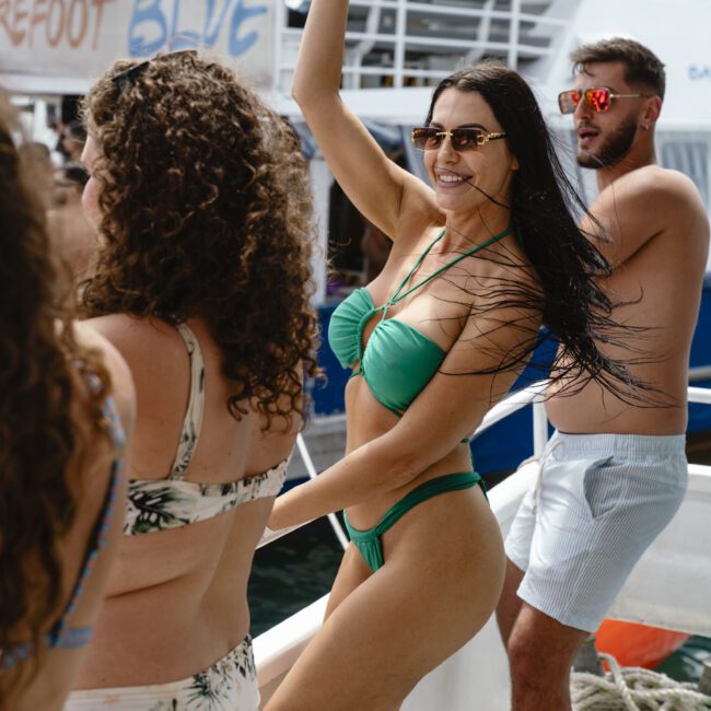 A group of people dancing and enjoying a boat party. A woman in a green bikini raises her arm joyfully. Other partygoers are around her, some wearing swimwear, with a boat named "Barefoot Blue" visible in the background.