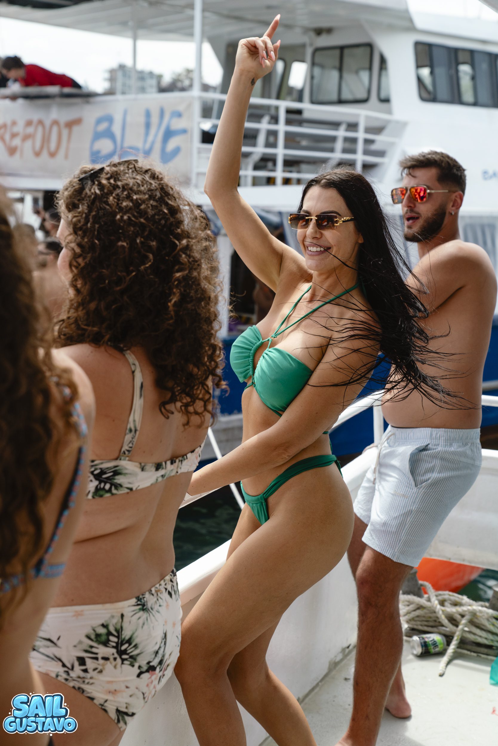 A group of people dancing and enjoying a boat party. A woman in a green bikini raises her arm joyfully. Other partygoers are around her, some wearing swimwear, with a boat named "Barefoot Blue" visible in the background.