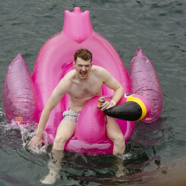 A person is happily floating on a pink inflatable in the shape of a chicken in a body of water. They're holding a toy resembling a drumstick and appear to be enjoying a drink.