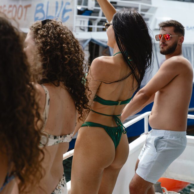 A group of people in swimwear enjoy a sunny day on a boat. A woman in a green bikini has an arm raised, while others are chatting and facing the sea. The background features a white boat.