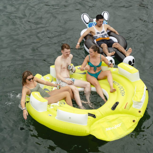 Four people are floating on an inflatable raft in water. They appear relaxed, with two seated on a yellow raft and the others on a black and white tube. The atmosphere is casual, suggesting they are enjoying a day out on the water.