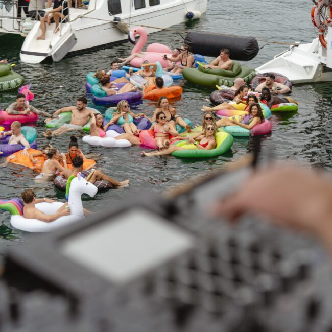 A group of people floating on colorful inflatable rafts, including a unicorn and flamingo, enjoy a party in the water near docked boats. A DJ setup is visible in the foreground, suggesting music at the event.