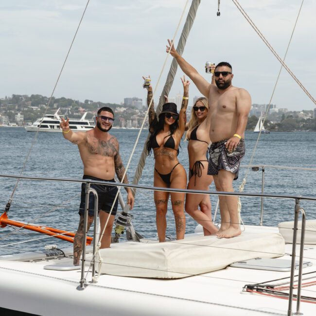 Four people in swimwear stand on a sailboat, smiling and holding drinks, with a scenic view of water and a distant city skyline in the background. They're making peace signs with their hands, enjoying a sunny day on the water.