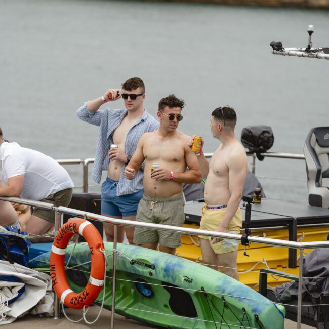 A group of men in casual summer attire stand on a boat by the water. Some hold drinks, and inflatable toys and life preservers are visible. The boat has a canopy and the water stretches into the background.