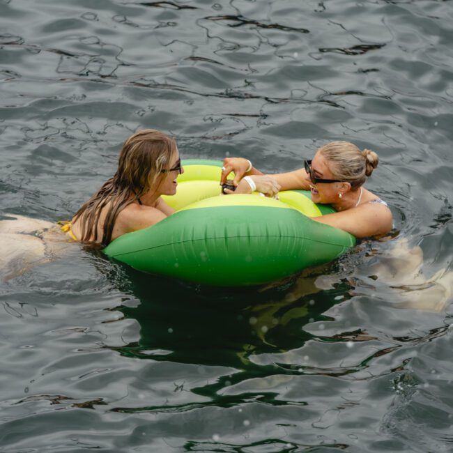 Two people are floating on a green inflatable in a body of water. They are smiling and appear to be enjoying themselves.