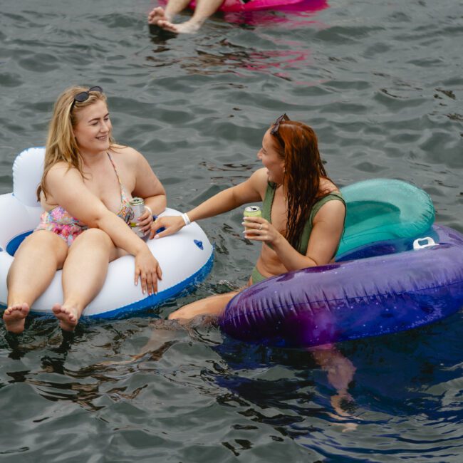 Two people are relaxing on inflatable tubes in the water. One, in a colorful bikini, sits on a white float, while the other, in a patterned swimsuit, sits on a purple float, holding a drink. Others float nearby in the background.