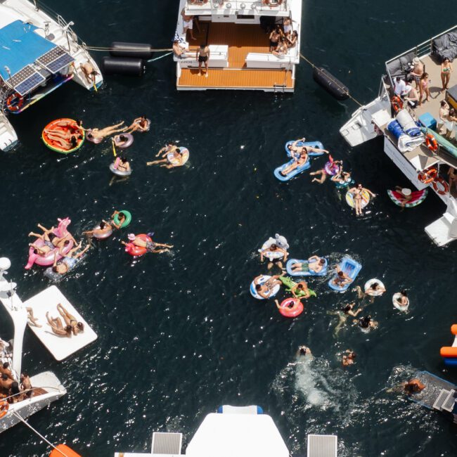 Aerial view of a lively boat party with people swimming and relaxing on colorful inflatables in the water. Several boats are anchored close together, and groups of people are gathered on deck enjoying the sunny day.