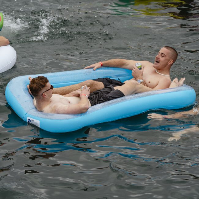 Three men enjoying a day at the water: two float on a blue inflatable raft, one holding a drink, while a third swims nearby. Another person is partially visible on a unicorn float. The scene is lively and relaxed.