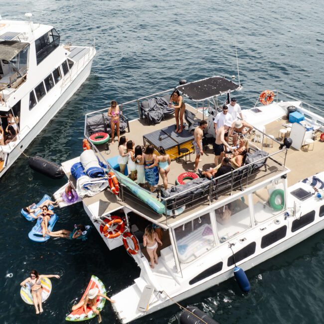 Aerial view of two boats on the water, crowded with people enjoying a sunny day. Some are standing on the decks while others relax on floats beside the boats. The water is calm and several life buoys are visible on the boat railings.