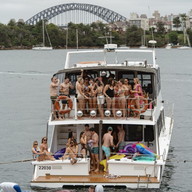 A large group of people are gathered on a boat having a party. Some are sitting, and others are standing and talking. The boat is on water with an iconic bridge and buildings visible in the background. Inflatable pool toys float nearby.