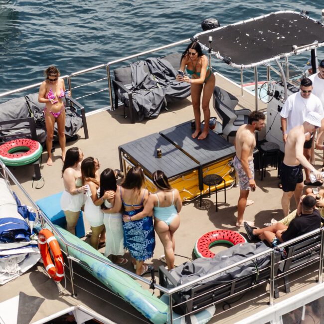 A group of people enjoys a sunny day on a boat deck, with some standing and others seated. Various supplies and life rings are visible. The surrounding water is calm, and everyone appears relaxed and social.