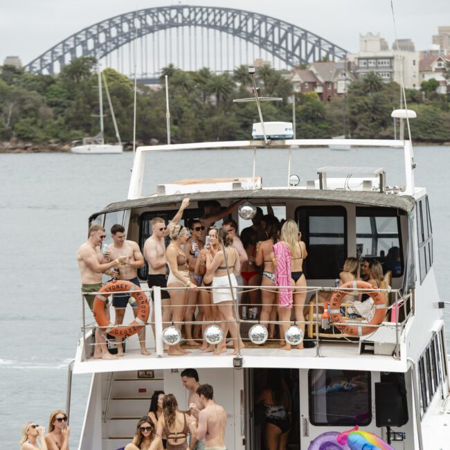 A group of people enjoying a party on a boat in a harbor. The Sydney Harbour Bridge is visible in the background, and people are socializing and taking photos on the upper deck of the boat.