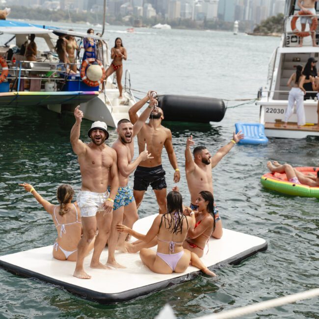 A group of people in swimsuits having fun on a floating platform in the water, with boats and more people in the background. Some are sitting, some are standing, and they're celebrating under a sunny sky.