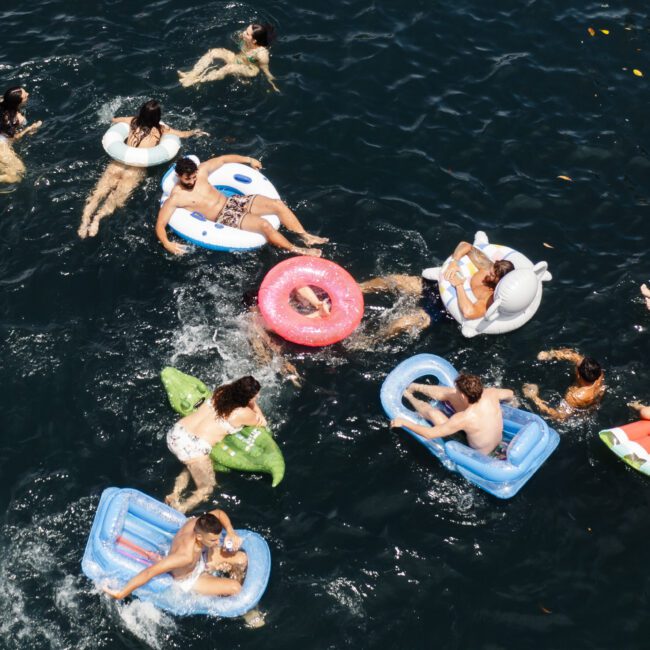 Aerial view of people swimming and floating on colorful inflatables in a dark blue body of water. The scene includes various float shapes, like rings and loungers, with individuals relaxing and swimming around.