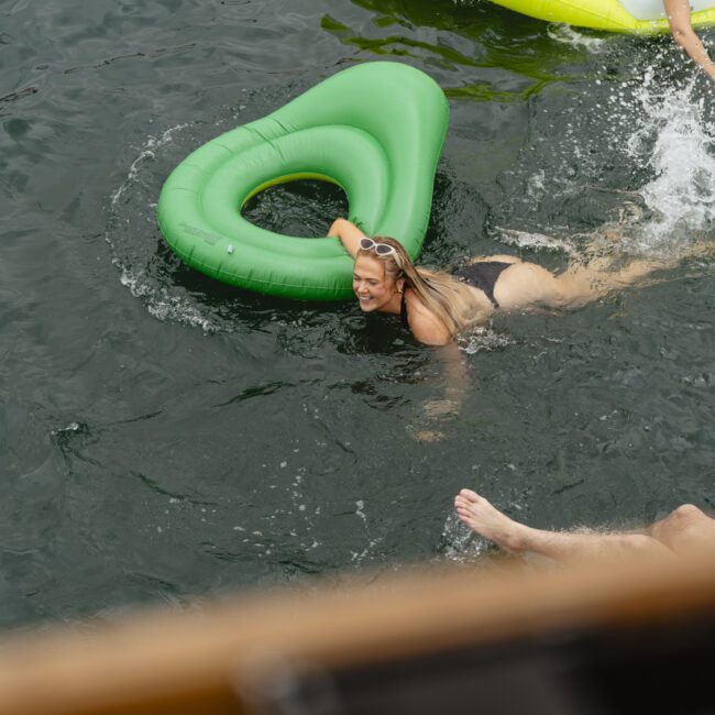 A person swims in a lake, smiling and holding onto a green, heart-shaped float. Nearby, a blue float and another person's leg are partially visible. The water is dark and slightly rippled.