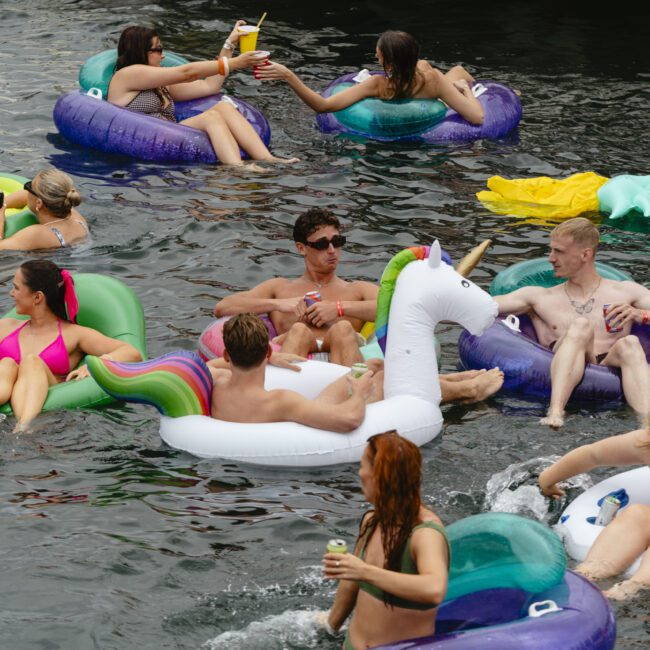 A group of people enjoying a sunny day on the water, lounging on colorful inflatable floats. Among them is a unicorn float. They are socializing, wearing swimsuits, and holding drinks. The scene is festive and lively.