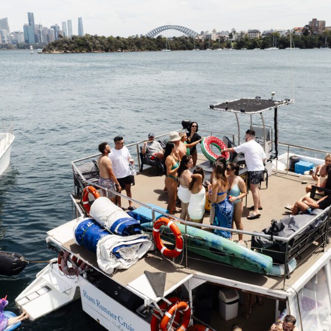 A group of people enjoying a party on a boat in a harbor. They are dancing and holding drinks on the deck. The skyline and a bridge are visible in the background. Other boats are on the water nearby.