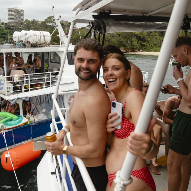 A man and a woman smiling on a boat deck, both in swimwear. The woman holds a phone, and the man holds a drink. In the background, other people are on a nearby boat with greenery and water visible.